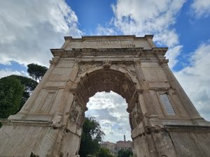 A low-angle view of the Arch of Titus, located in the Roman Forum (Rome, Italy). The ancient Roman arch is made of stone and features intricate carvings and details.