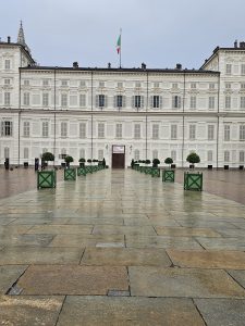 The Royal Palace of Turin, also known as Palazzo Reale, is on a rainy and cloudy day. The palace, a large, white building with many windows and closed shutters, dominates the background. A flag flies atop the center of the palace. In the foreground, a stone-paved courtyard stretches out.