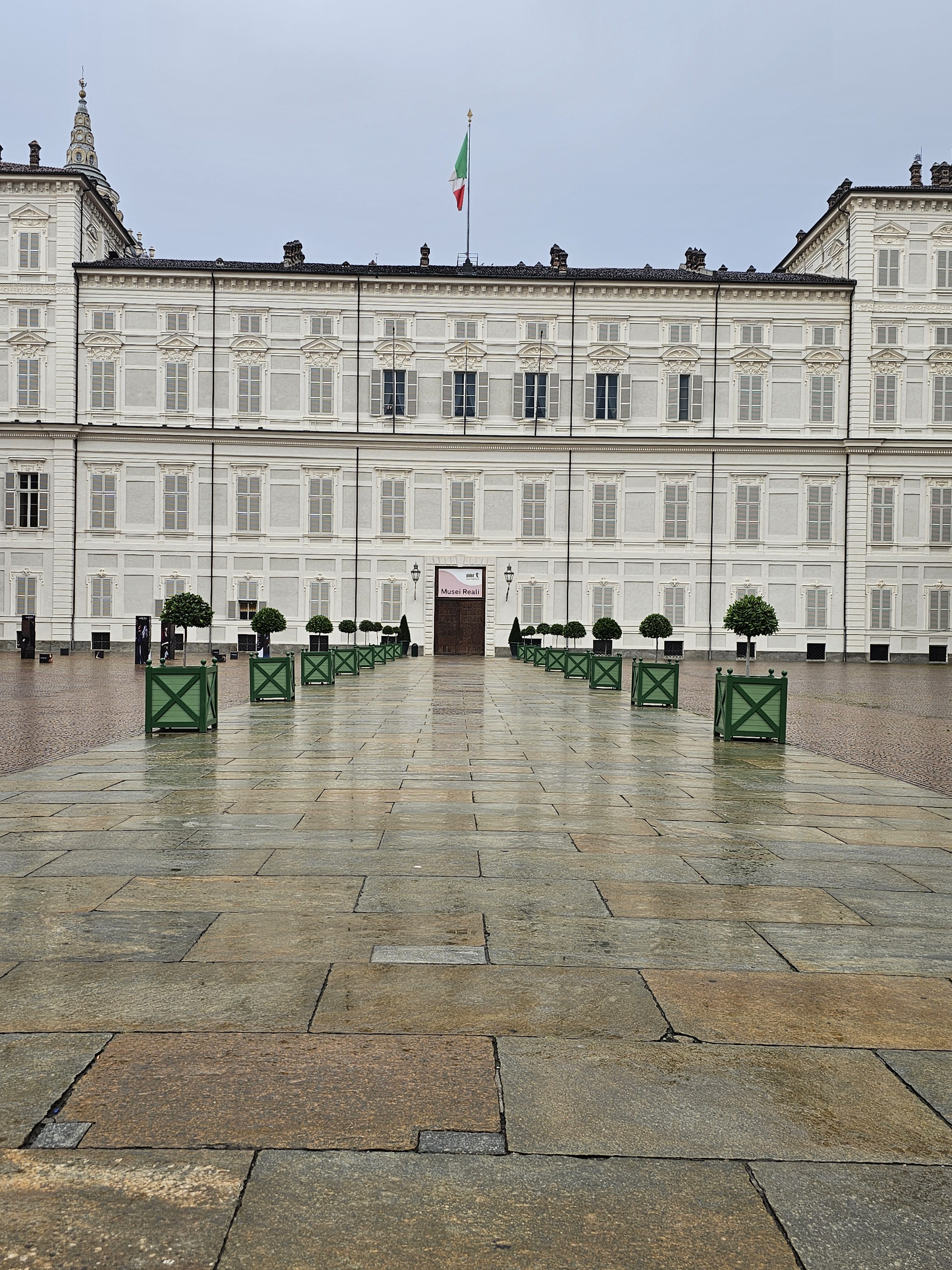 The Royal Palace of Turin, also known as Palazzo Reale, is on a rainy and cloudy day. The palace, a large, white building with many windows and closed shutters, dominates the background. A flag flies atop the center of the palace. In the foreground, a stone-paved courtyard stretches out.