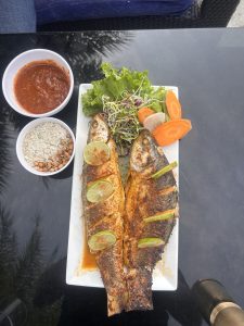 Grilled whole fish served on a white rectangular plate, garnished with lime slices and accompanied by a side of fresh lettuce, sliced radishes, carrots, and a small salad.