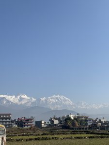 A cityscape with multi-story buildings in the foreground, set against a backdrop of majestic snow-capped mountains under a clear blue sky.
