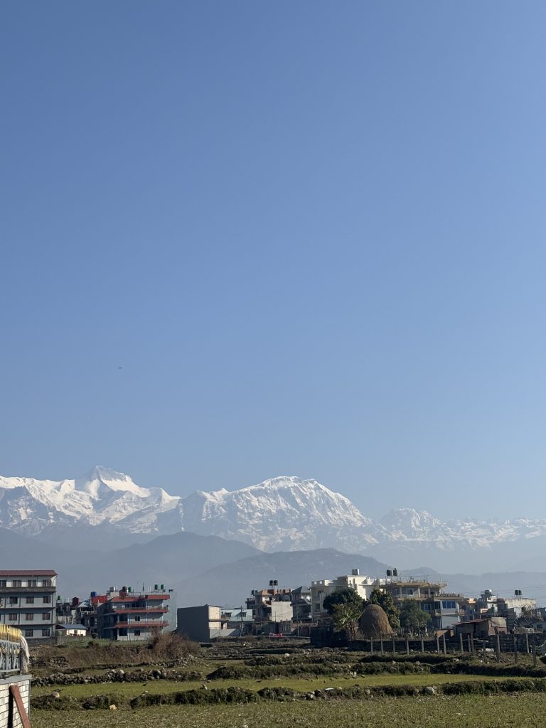 A cityscape with multi-story buildings in the foreground, set against a backdrop of majestic snow-capped mountains under a clear blue sky.