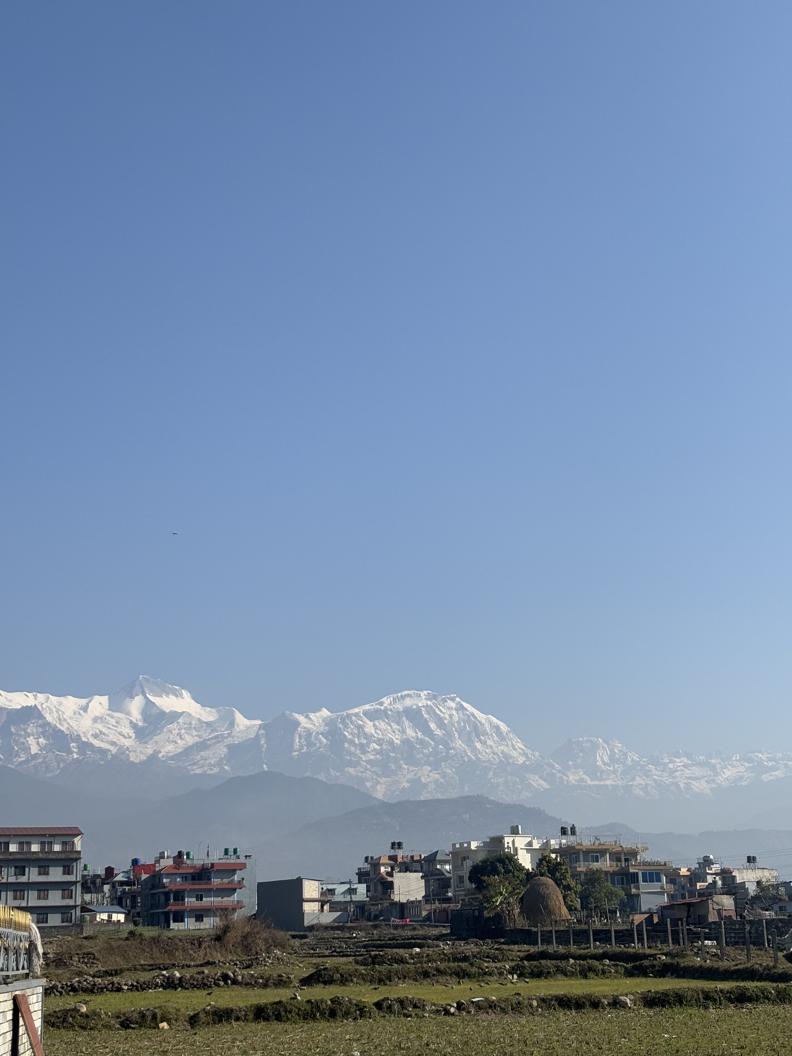 A cityscape with multi-story buildings in the foreground, set against a backdrop of majestic snow-capped mountains under a clear blue sky.