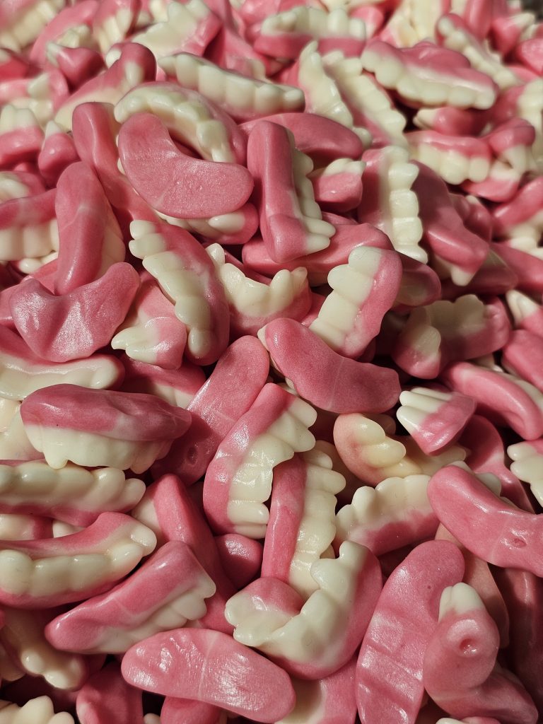 A close-up of a pile of pink and white gummy candies shaped like teeth. The pink part of the candies represents the gums, while the white part is shaped like teeth with detailed ridges.