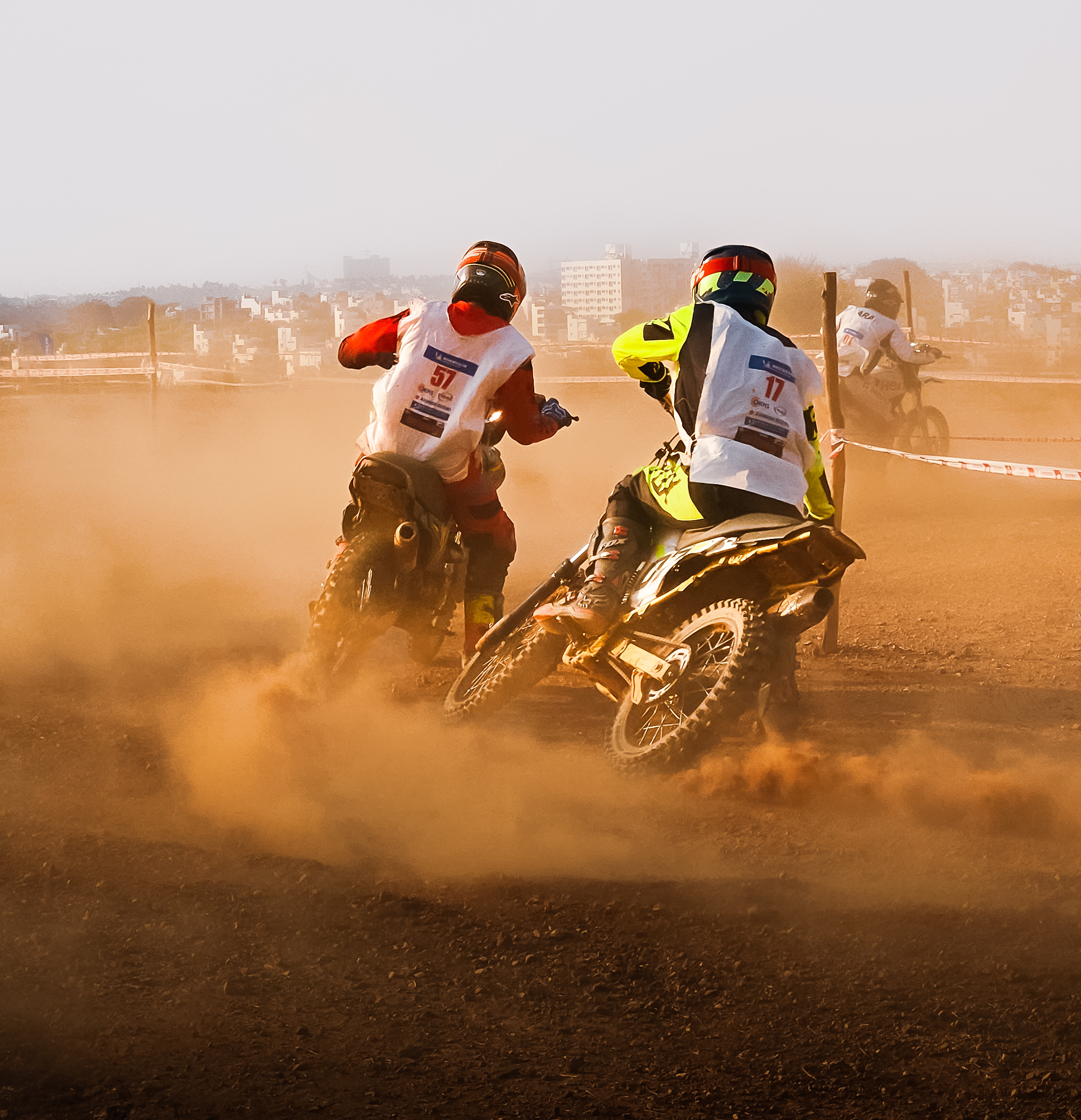 Two motocross riders in protective gear race around a dirt track, kicking up dust as they navigate a turn. Other riders can be seen in the background, with a cityscape in the distance under a clear sky.
