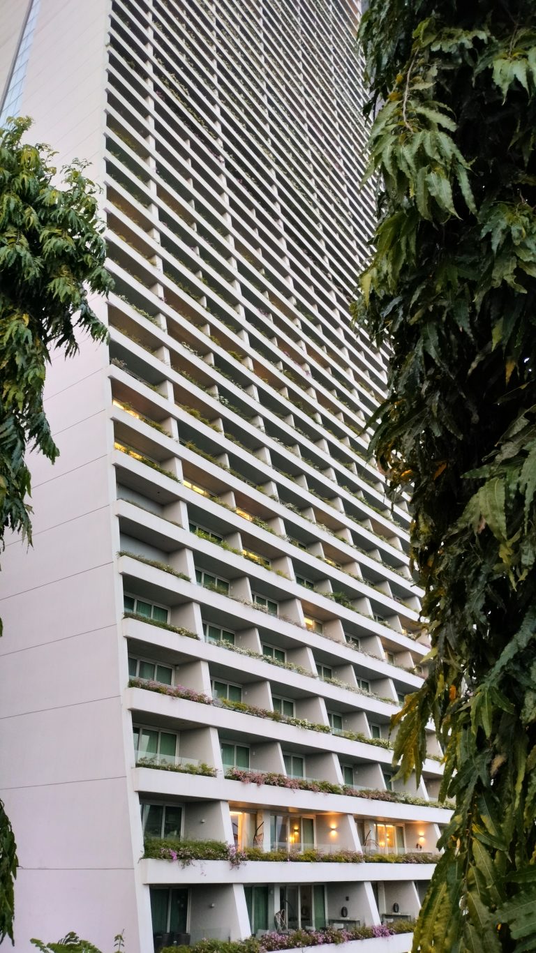 A tall building with a modern architectural design characterized by rows of balconies filled with greenery. Tall trees frame the sides of the building.