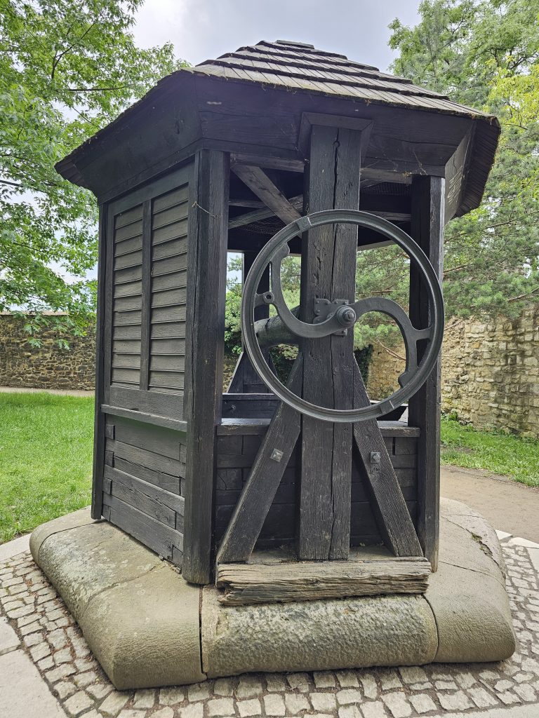 A traditional wooden well structure with a roof, a crank wheel, and a stone base, located in Vy?ehrad, Prague.