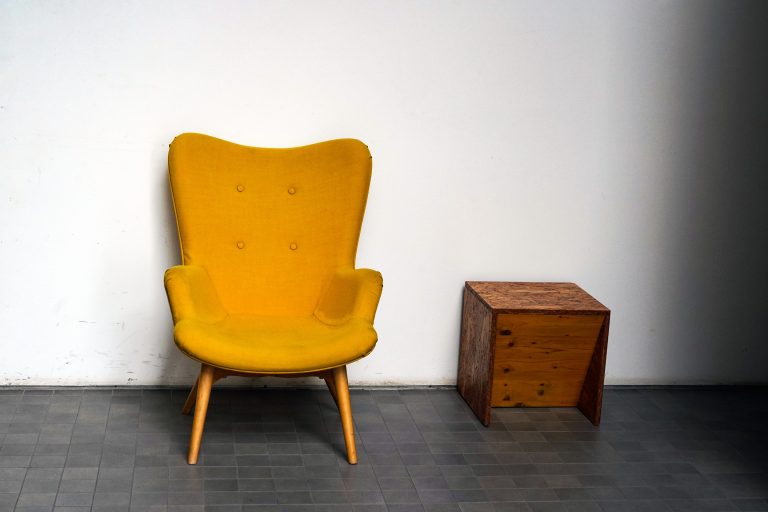 A yellow upholstered armchair with wooden legs is placed next to a small wooden cube-shaped side table. The setting is against a plain white wall on a tiled floor.