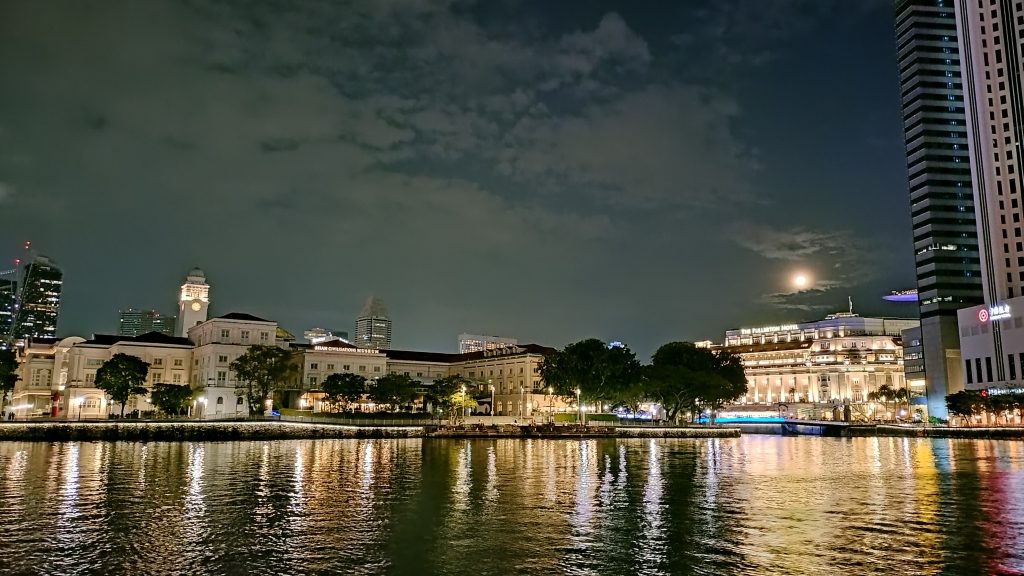 A serene night view of Singapore’s waterfront, showcasing the beautifully illuminated Asian Civilisations Museum and the iconic Fullerton Hotel. The calm waters reflect the warm lights from the historic buildings and surrounding structures, while the full moon peeks through scattered clouds, adding a touch of tranquility to the vibrant cityscape.