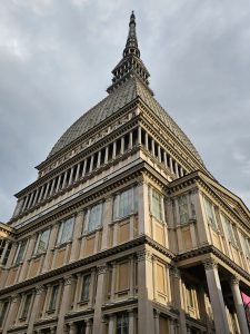 A low-angle view of the Mole Antonelliana in Turin, Italy, featuring its distinctive tall spire and neoclassical architectural details. The building's facade is captured against a cloudy sky, emphasizing its height and grandeur.
