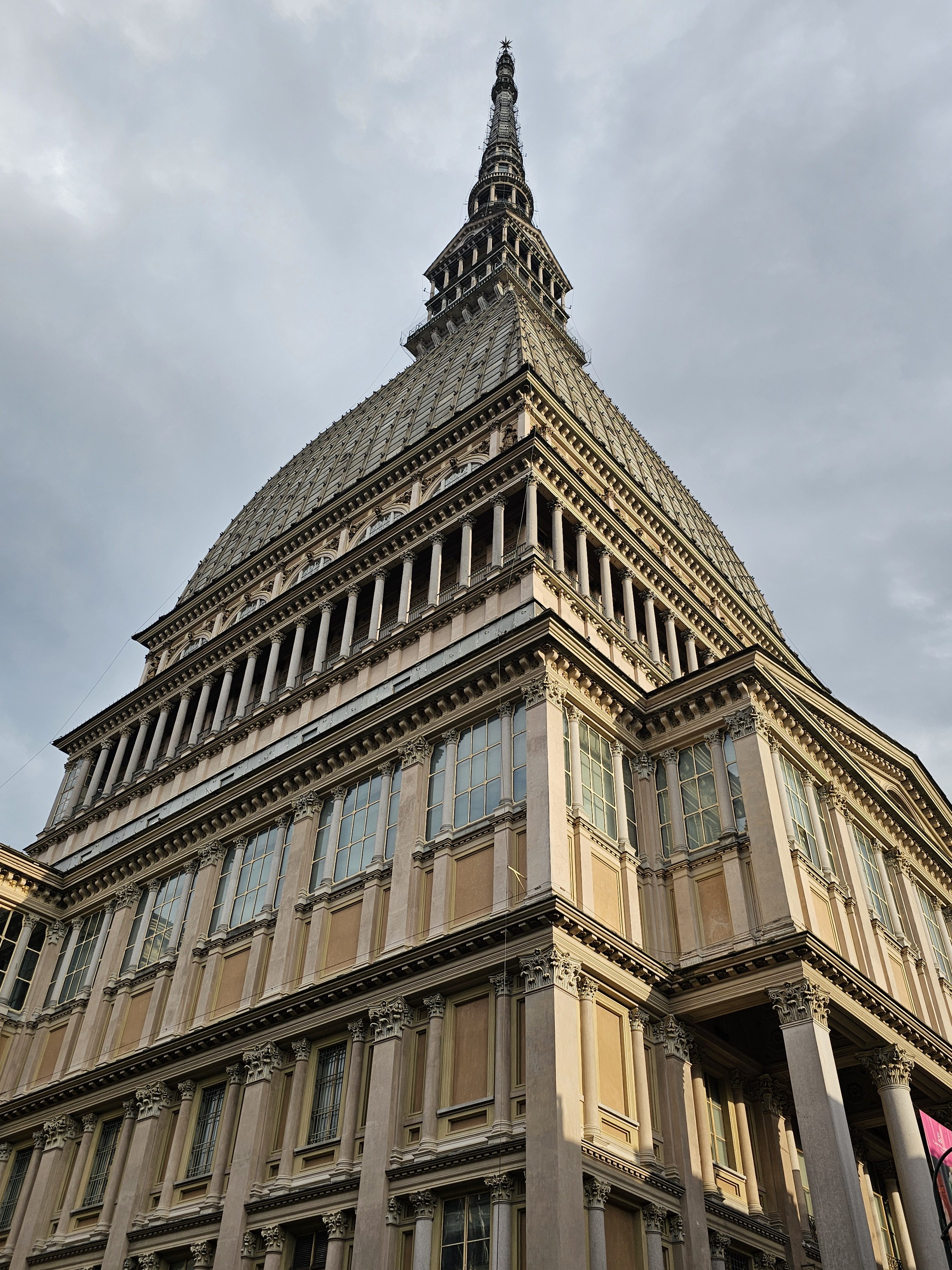 A low-angle view of the Mole Antonelliana in Turin, Italy, featuring its distinctive tall spire and neoclassical architectural details. The building's facade is captured against a cloudy sky, emphasizing its height and grandeur.