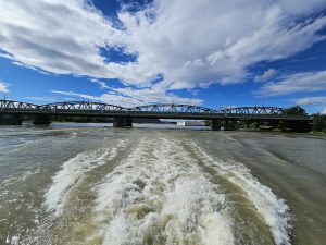 A view from the back of a boat moving on Danube river, with a truss bridge spanning over the river in the background, under a partly cloudy sky. The wake of the boat is visible in the foreground. Captured from Vienna, Austria. 
