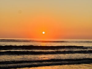 Sunset over the ocean with the sun casting a warm glow across the water and gentle waves rippling towards the shore.
