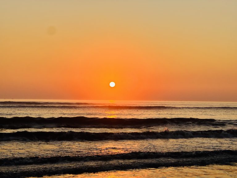 Sunset over the ocean with the sun casting a warm glow across the water and gentle waves rippling towards the shore.