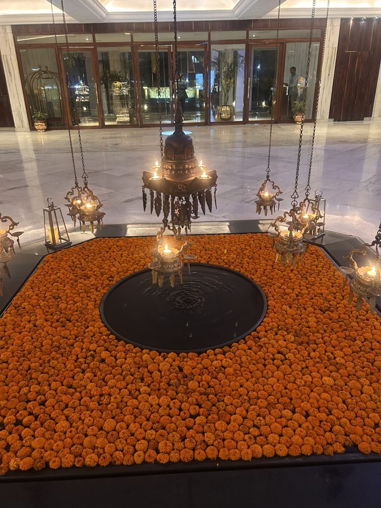 A decorative indoor installation featuring a large square base covered with orange marigold flowers, centered around a circular water feature. Elegant metal lamps with lit candles are suspended from above, casting a warm glow. In the background, there are glass doors and a spacious marble floor.