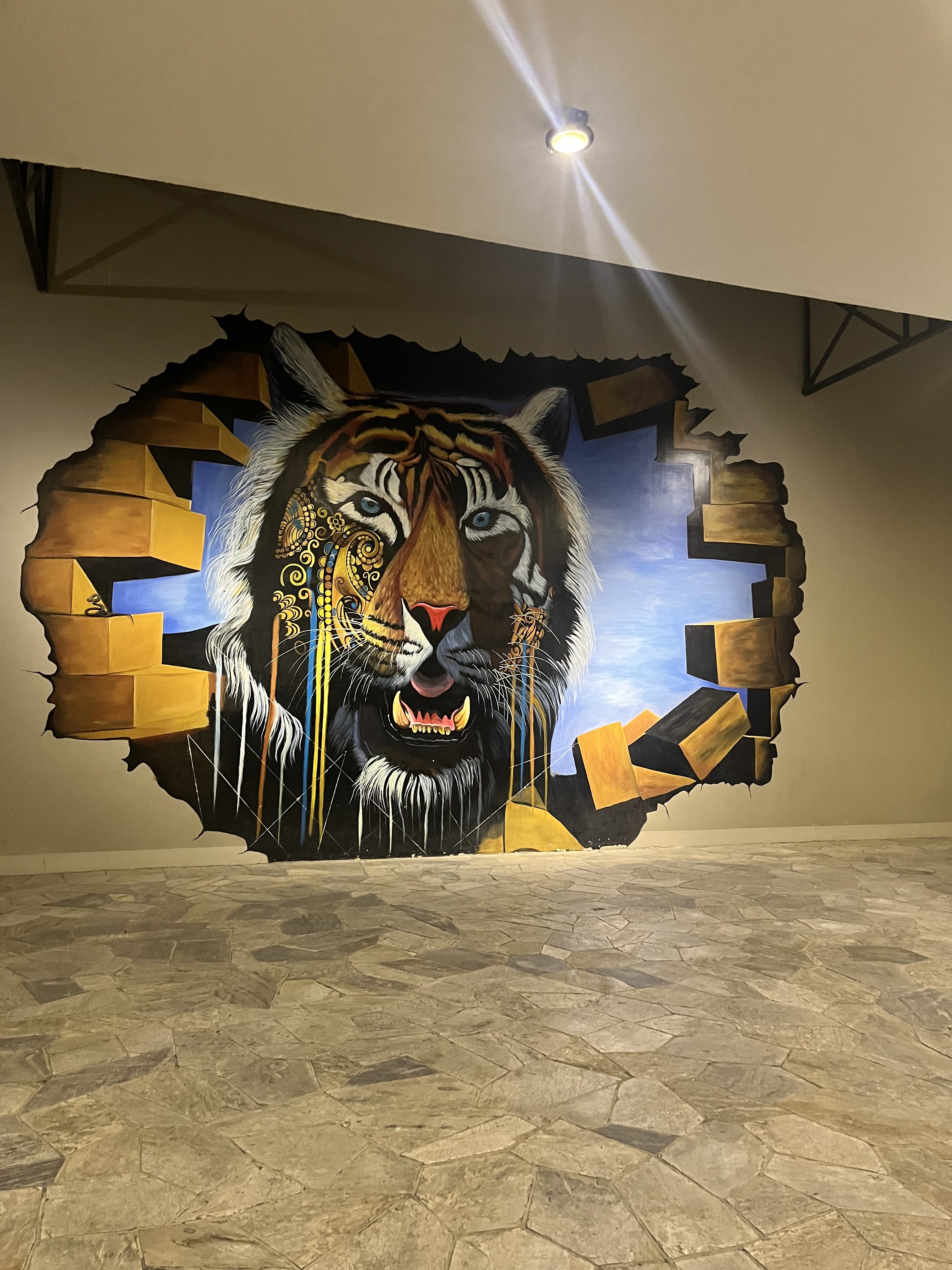 A mural painting of a tiger's face on a wall, giving the illusion that it is breaking through the surface. The tiger features detailed eyes and prominent teeth, and one side is adorned with intricate golden patterns. The background is a mix of a blue sky and golden geometric shapes. The scene is illuminated by a spotlight above.