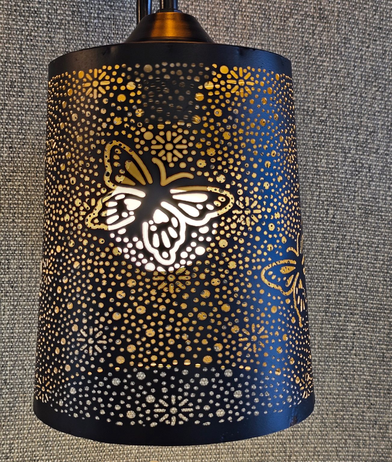 A black metal lantern with intricate butterfly and flower cutouts casts a decorative glow through its perforations onto a textured gray background. It was spotted in an electrical shop in Kozhikode, Kerala.