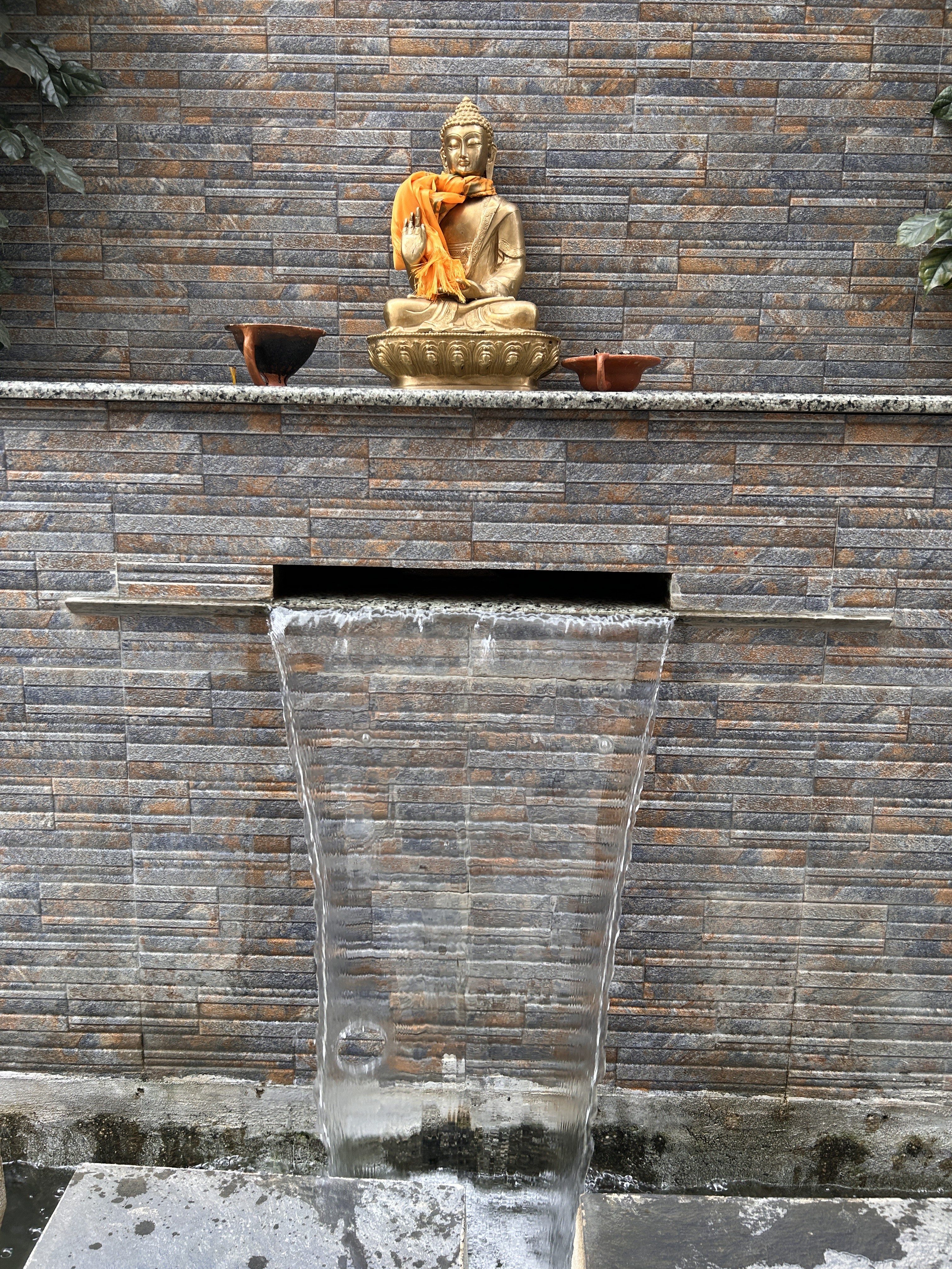 A golden Buddha statue draped with an orange cloth sits on a ledge against a textured stone wall. Two small vessels flank the statue. Below, water cascades from an opening in the wall into a small pool.