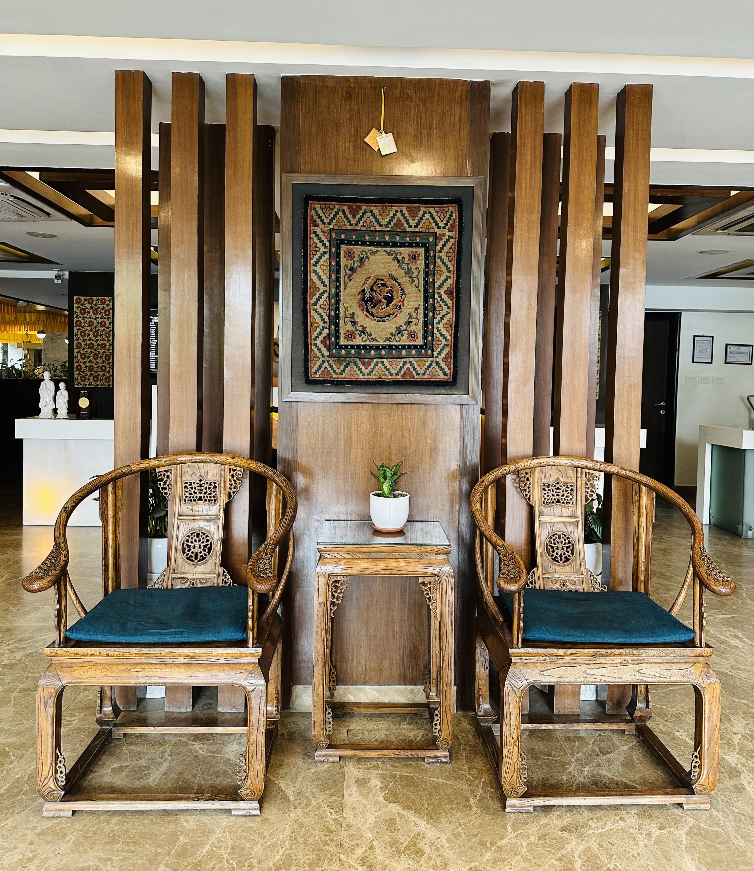Elegant wooden chairs with intricate carvings are positioned on either side of a small wooden table holding a potted plant.