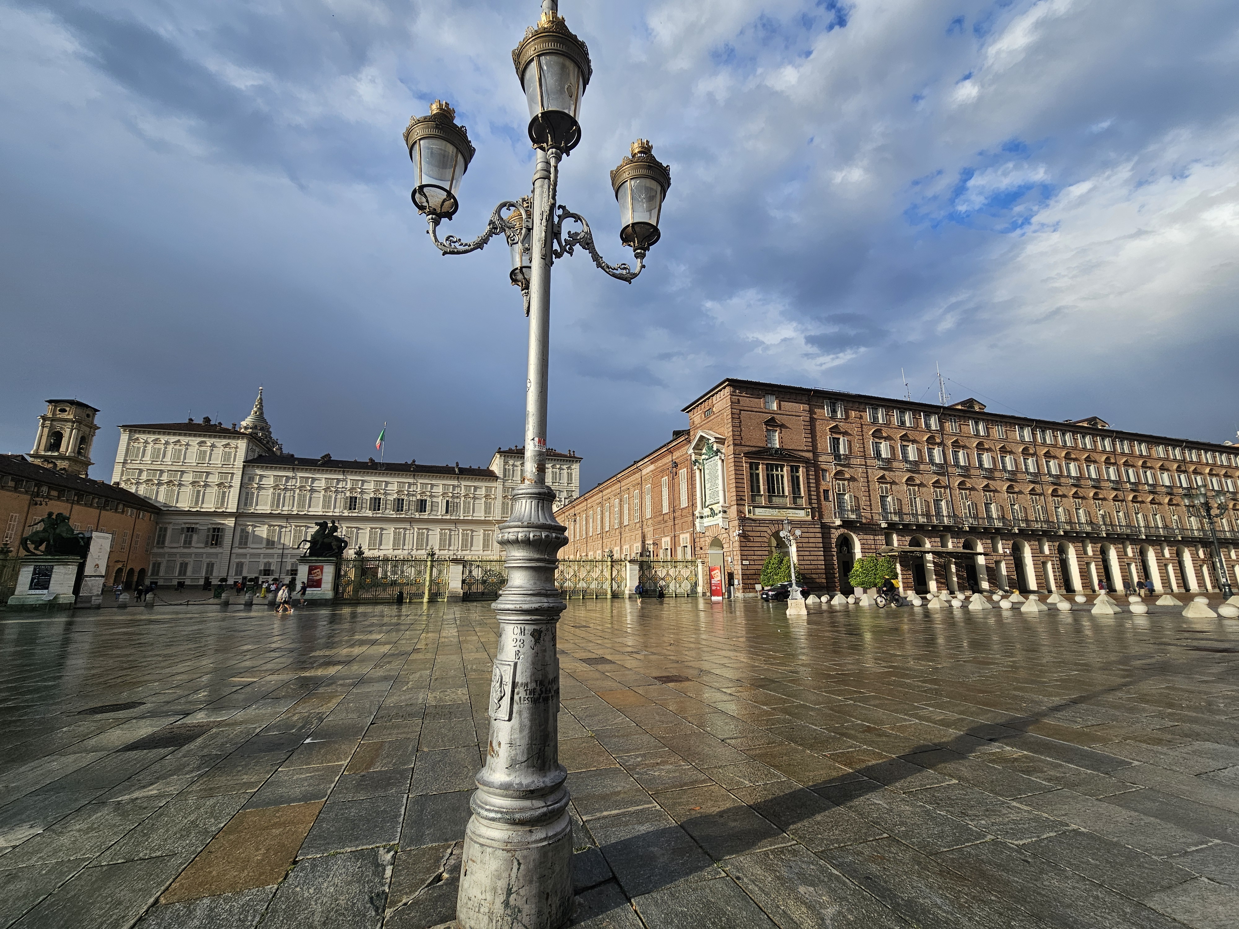 The Piazza Castello in Turin, Italy, features the Royal Palace of Turin and Palazzo Madama under a cloudy sky. A prominent lamppost stands in the foreground and the architecture of the buildings reflects a blend of Baroque and earlier styles, characteristic of Turin's historical center.
