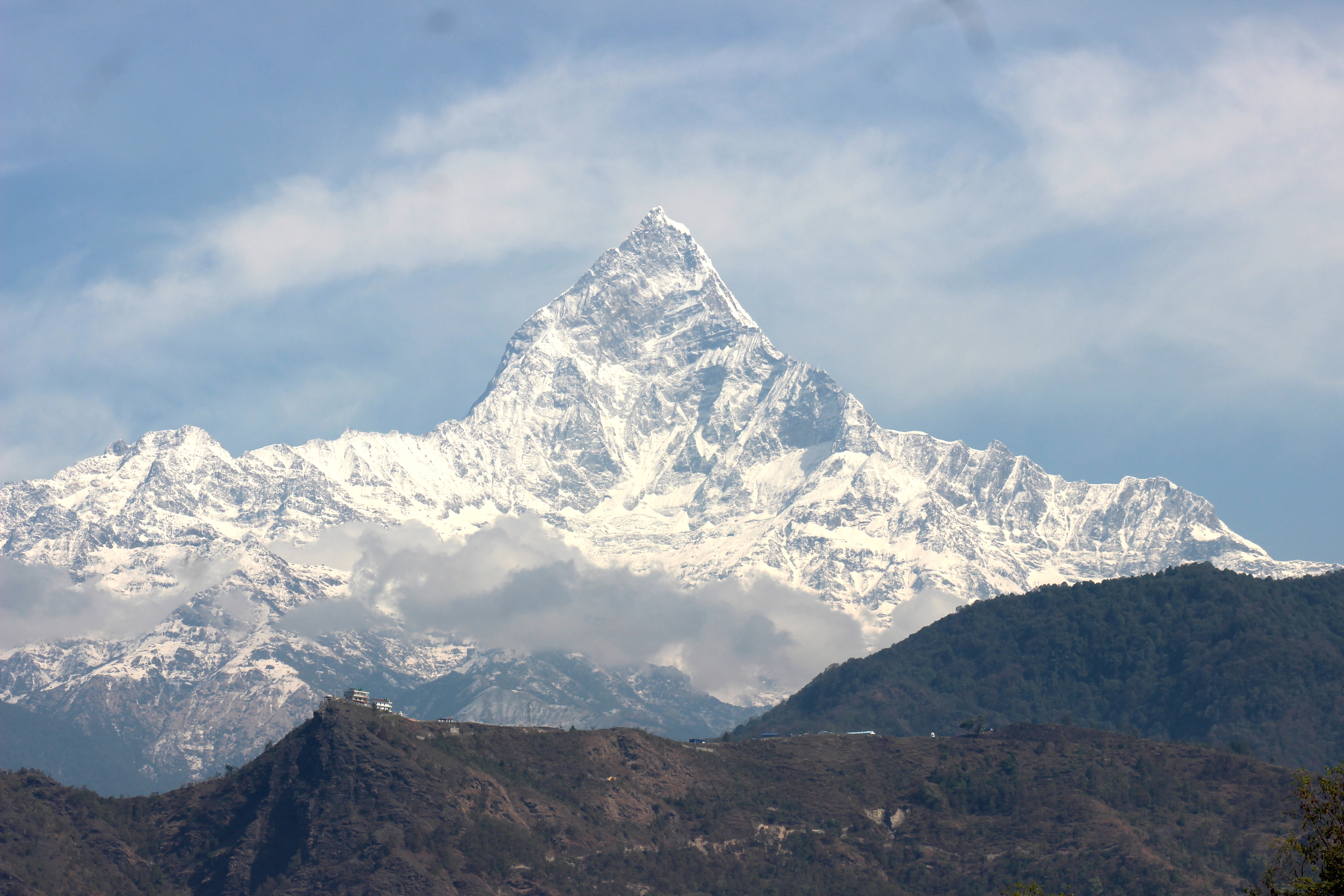 A majestic snow-covered Fishtail mountain peak stands prominently against a blue sky, with lower rocky hills in the foreground partially covered by vegetation and clouds.