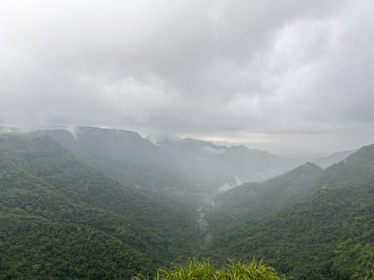 A scenic view of lush green mountains shrouded in mist and clouds, with a foggy atmosphere under a grey, overcast sky.