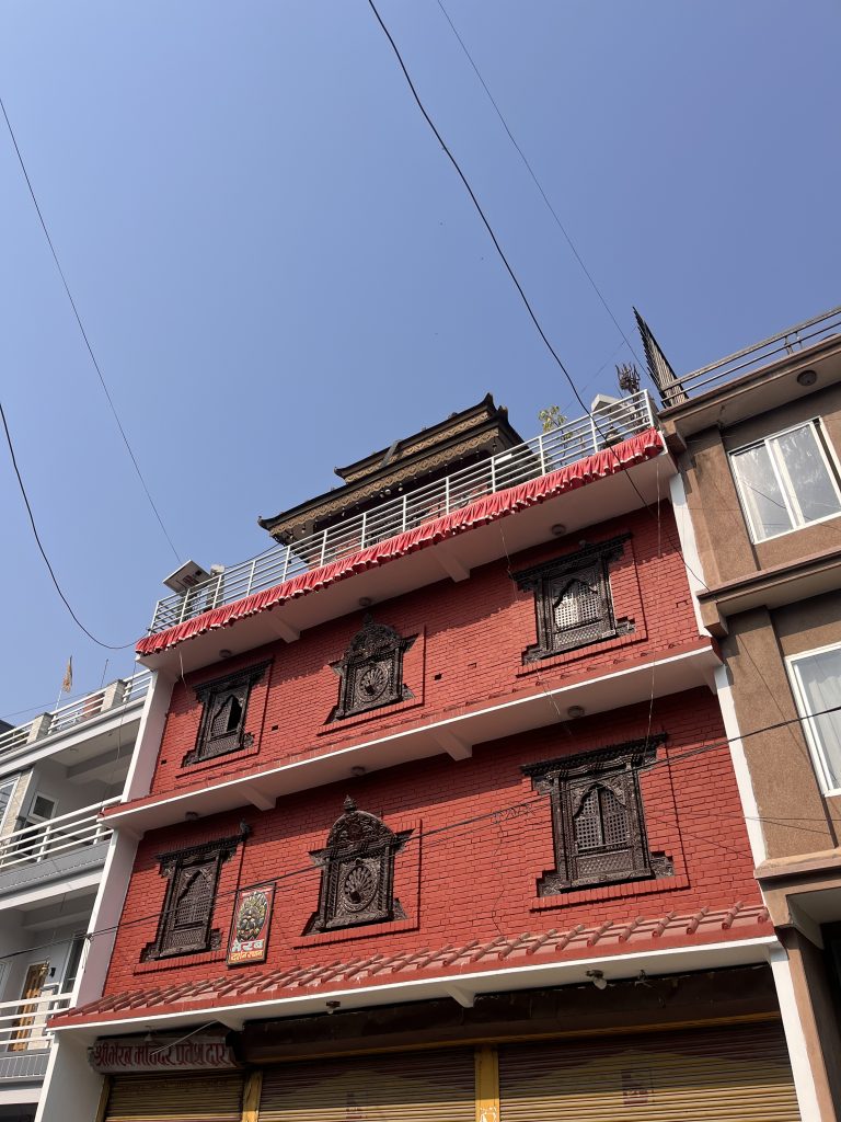 A multi-story building with red brick facade featuring ornate, dark decorative windows. A traditional style rooftop adorns the top of the structure. Neighboring buildings with balconies are visible, and the sky is clear blue overhead.