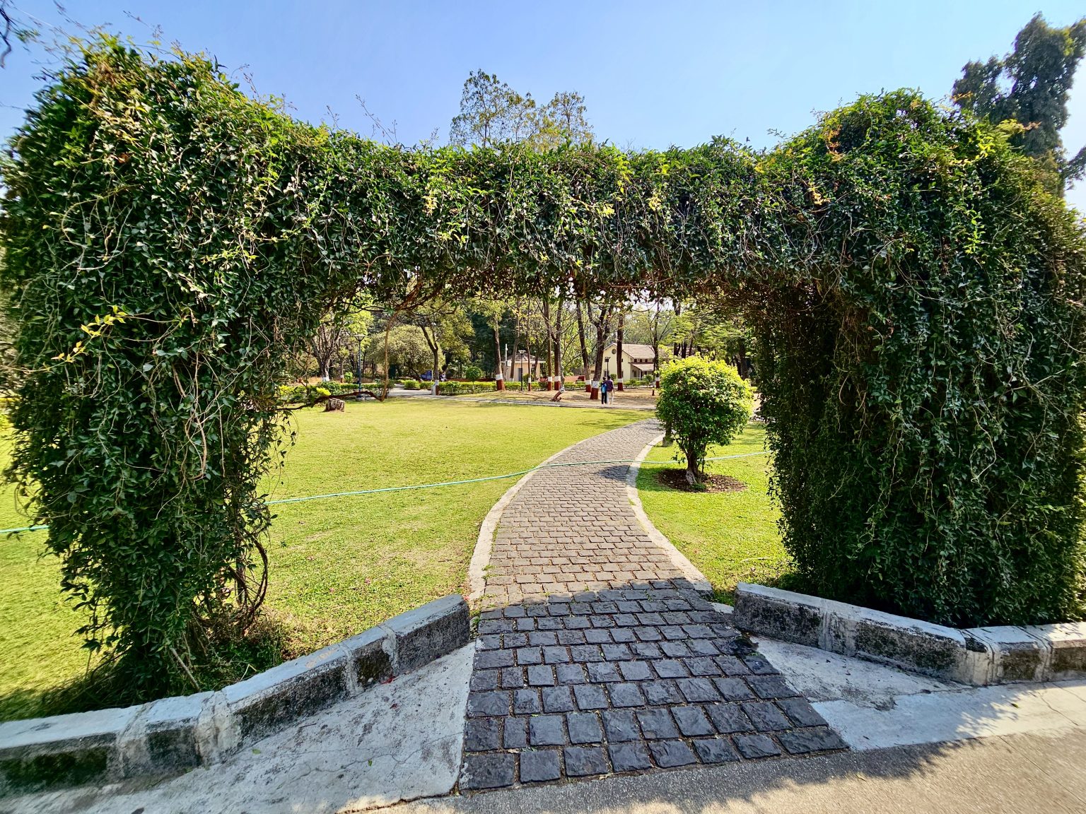 A scenic view of Aga Khan Palace Garden. Stone pathway, paved with dark grey stones, leads into the park, framed by a natural archway of thick green foliage. The archway is formed by dense bushes.