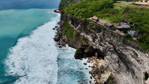 Aerial view of a cliffside restaurant south of Uluwatu Temple in Bali, perched above the turquoise ocean with waves crashing against the rocky shore.
