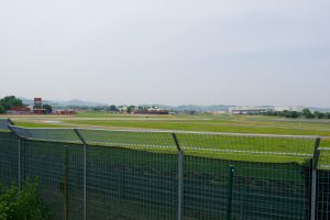 View of a racetrack surrounded by green grass and fencing, with buildings and hills visible in the distance under a cloudy sky.
