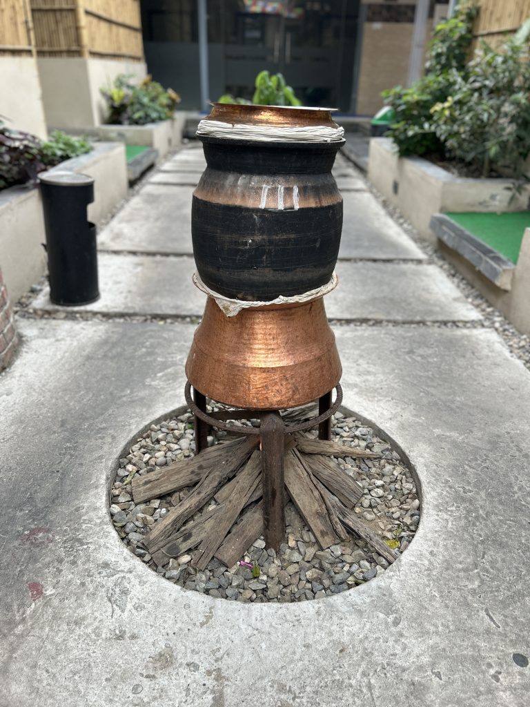 A traditional tandoor setup with stacked clay and copper pots, placed within a circular stone and gravel area. Wooden pieces are arranged around the base, resembling a firewood setup. The background features a path lined with plants and a bamboo fence.