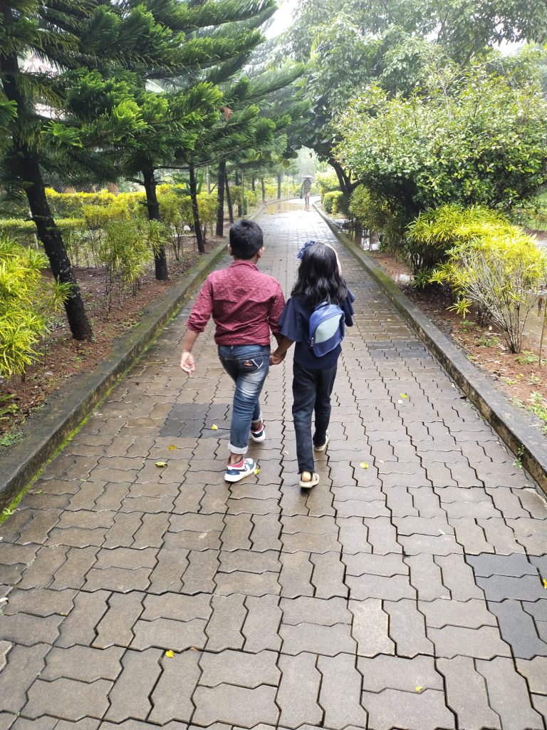 Two children walking hand in hand down a tree-lined brick path in a park, surrounded by lush greenery. They are dressed casually and one child is carrying a backpack.