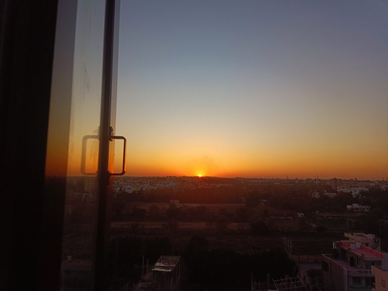 A breathtaking sunset view from a window, casting a golden glow over the cityscape and blending warm hues into the sky.