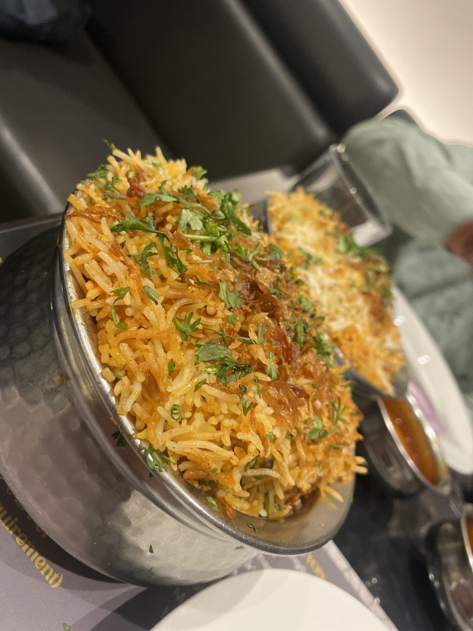 A close-up of a dish of biryani served in a metal bowl, garnished with herbs and fried onions. The background includes another bowl of biryani and a glass, partially visible.
