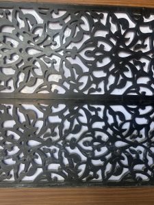 A dark, intricately carved wooden panel with an abstract floral pattern. The design allows light to filter through from behind, creating a contrast with the lighter background. The panel rests on a wooden surface.
