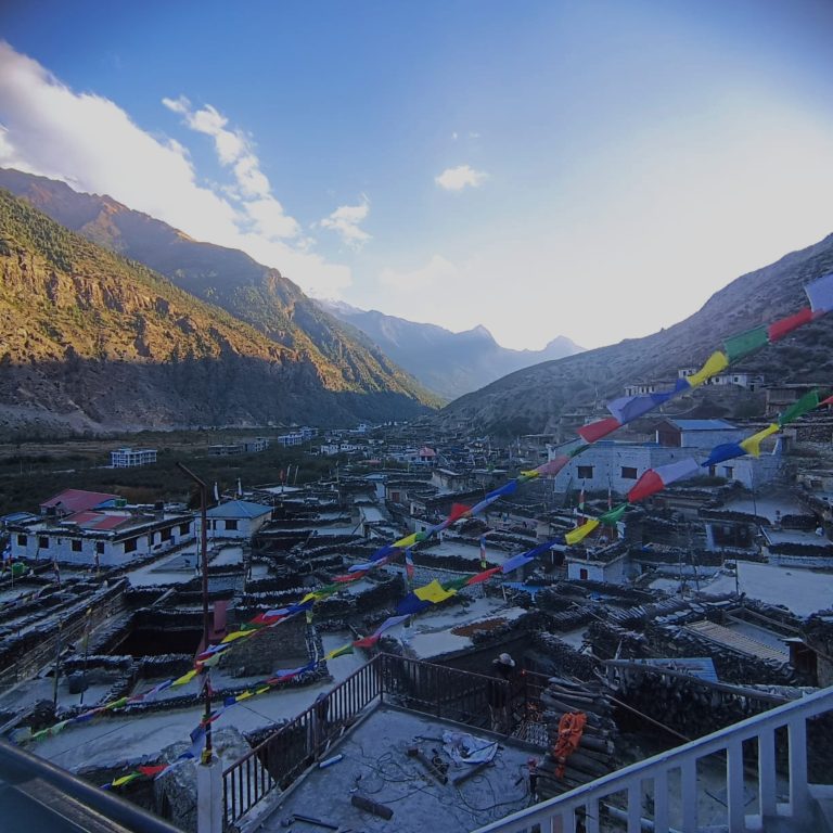 A mountain village nestled in a valley with colourful prayer flags in the foreground. The sunlit landscape shows rugged mountains and rooftops of traditional stone buildings. The photo taken at Marpha village, Nepal.