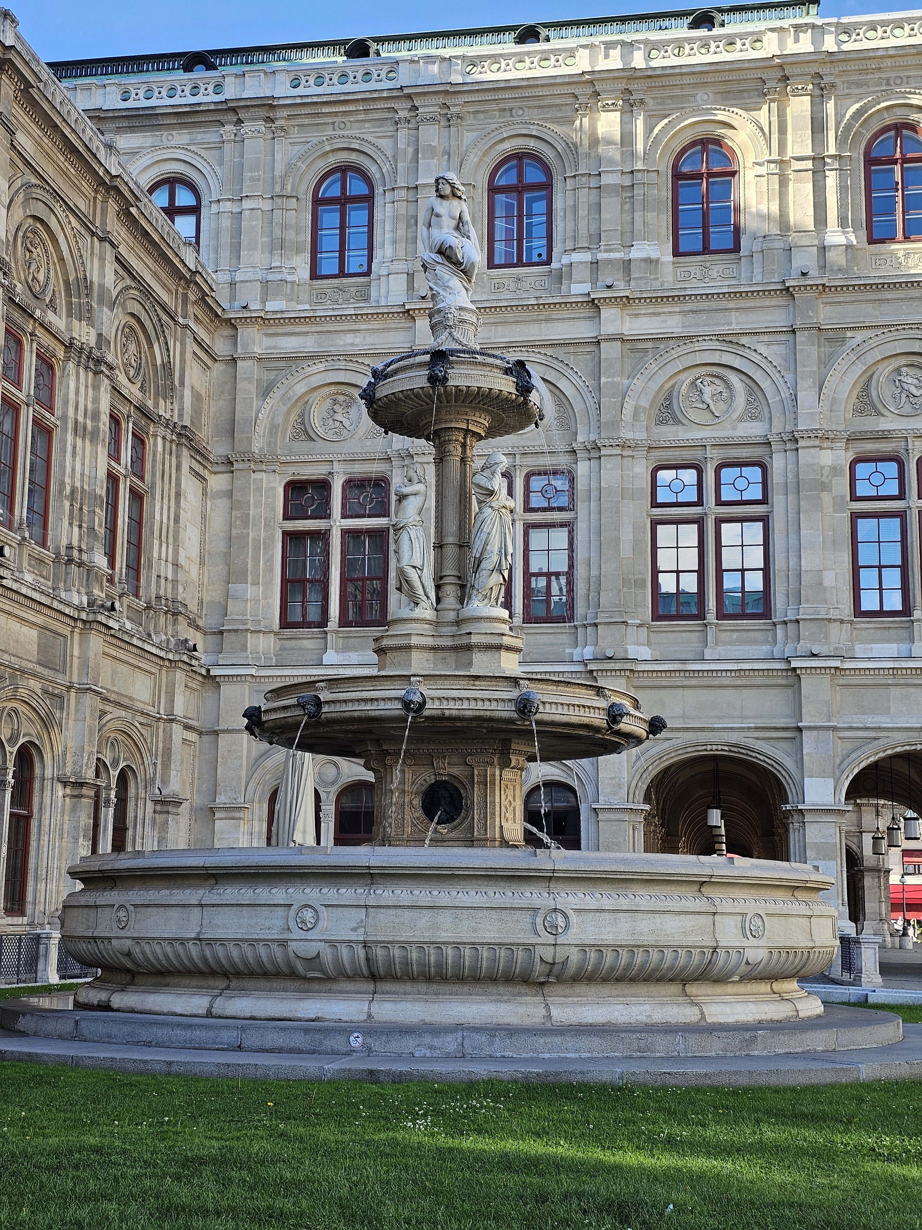 The Opernbrunnen fountain, located at the Vienna State Opera. The fountain features tiered basins and sculptures against the backdrop of the opera house's facade, characterized by arched windows and ornate details. The structure is made of stone, and a patch of green grass is visible in the foreground.