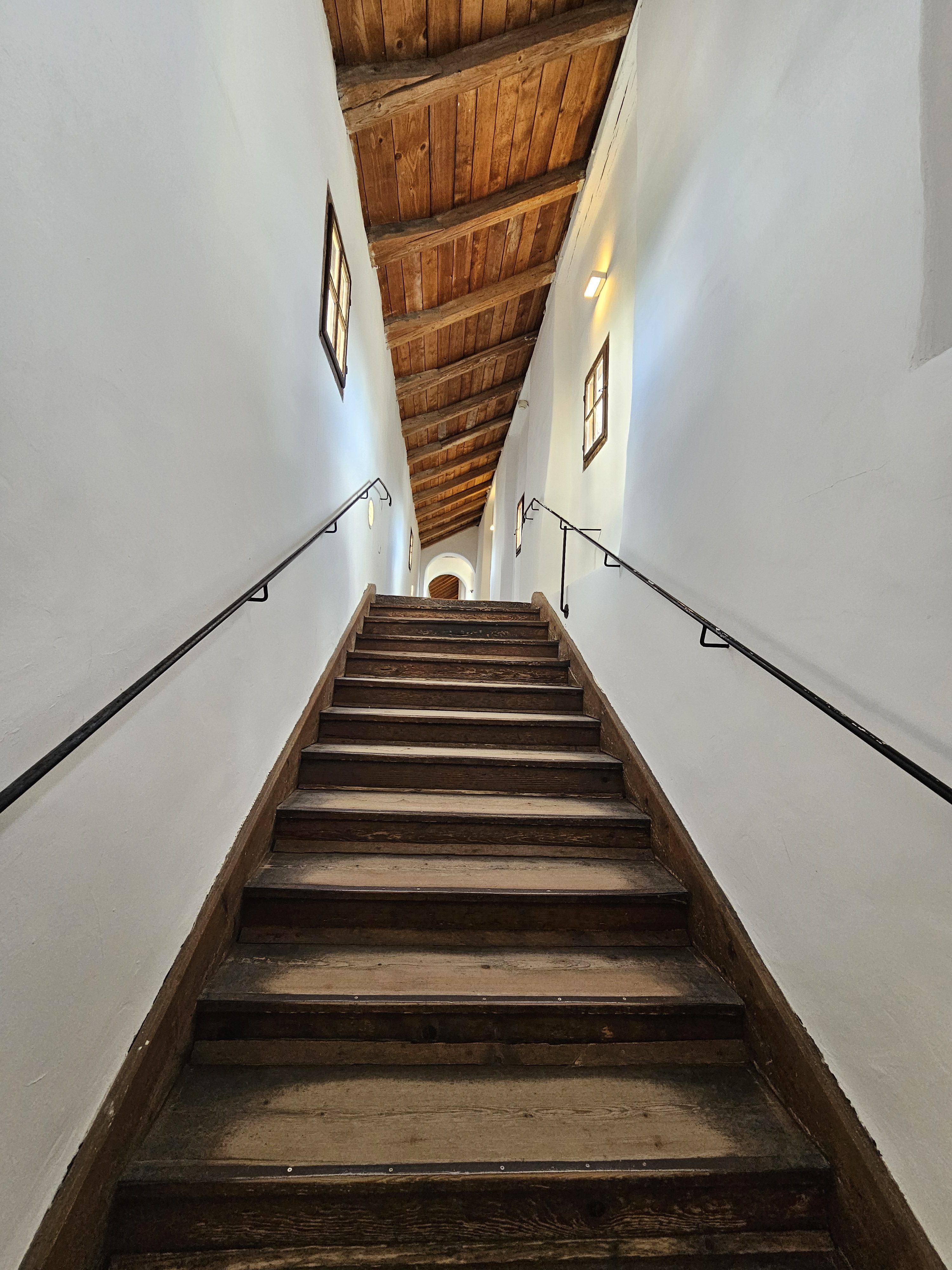 A wooden staircase ascending between white walls, featuring a dark wood ceiling and black metal handrails. Small windows are visible on the walls, and the steps are worn, indicating frequent use. Captured from Hohensalzburg Fortress, Salzburg, Austria.