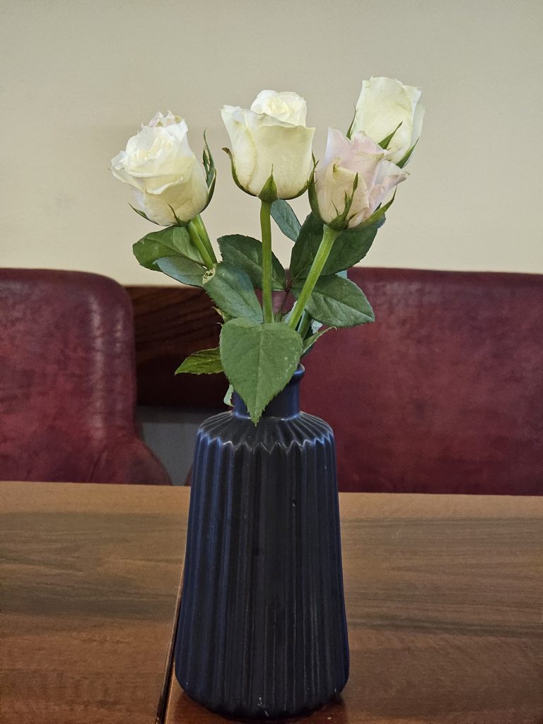 A dark blue, ridged vase holding four white roses with green leaves, set on a wooden table. Two burgundy-colored chairs are partially visible in the blurred background behind the vase