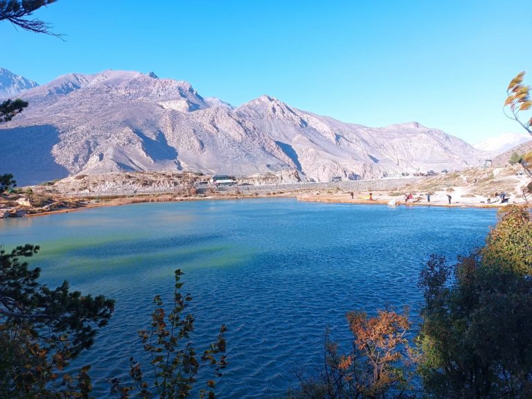 A scenic view of a tranquil lake surrounded by mountainous terrain under a clear blue sky. The lake reflects shades of green and blue, with some trees and vegetation along the shoreline. In the background, several people can be seen walking near the banks, and a road with a vehicle is visible along the hills.