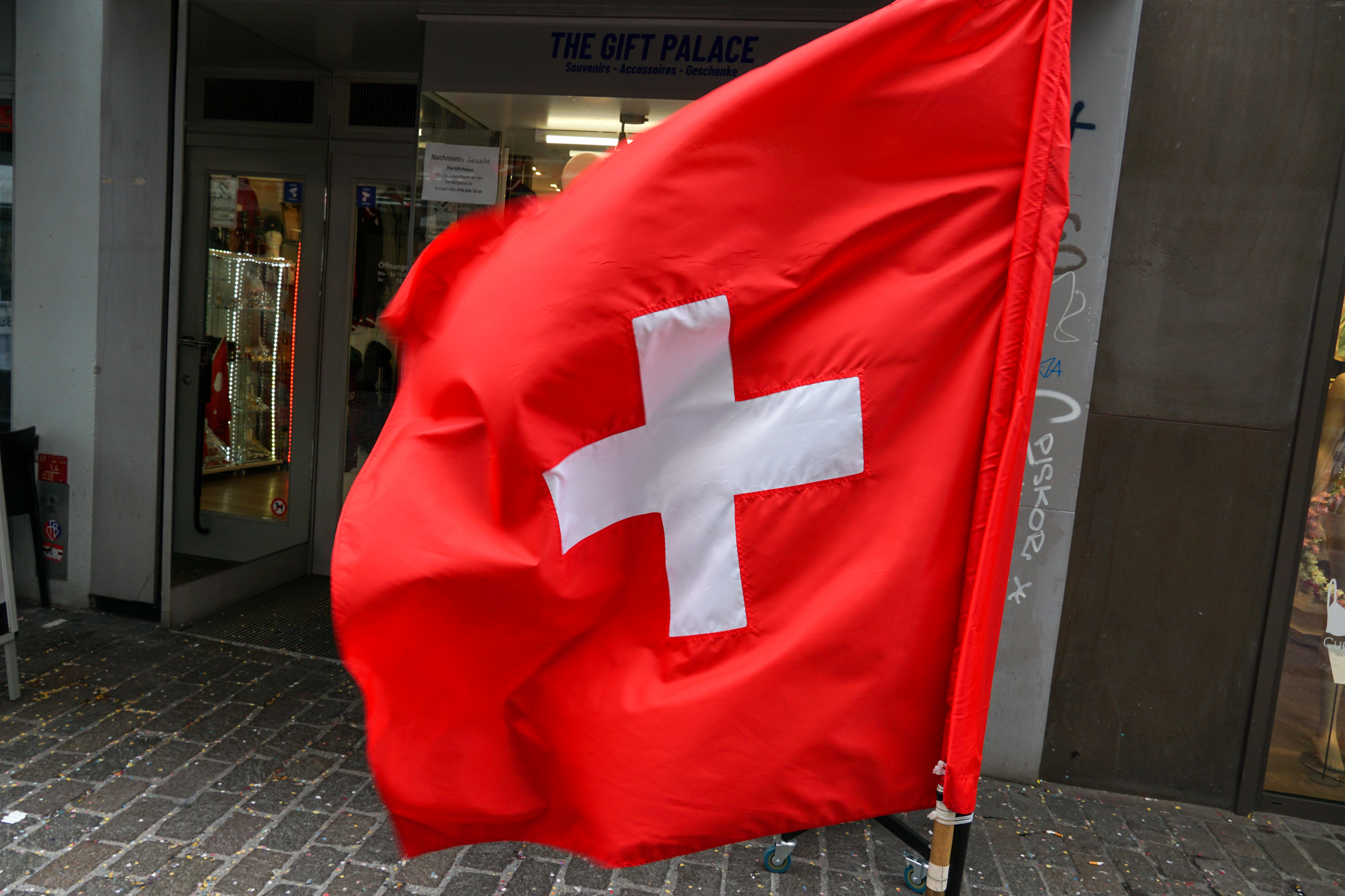 A large Swiss flag with a white cross on a red background is displayed prominently on a city street, in front of a shop entrance