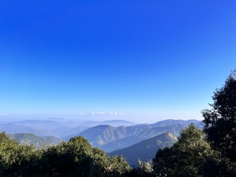 A scenic view of layered mountain ranges with green foliage in the foreground and snow-capped peaks visible in the distance under a clear blue sky.