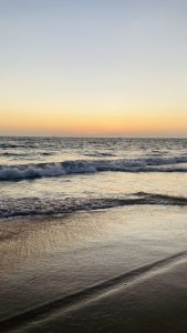 A serene beach at sunset with gentle waves rolling onto the shore, reflecting the warm hues of the sky in golden and orange tones.