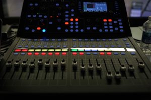 A close-up of a professional audio mixing console with various sliders, buttons, and a digital display screen, illuminated by colored lights. There is a water bottle to the side.