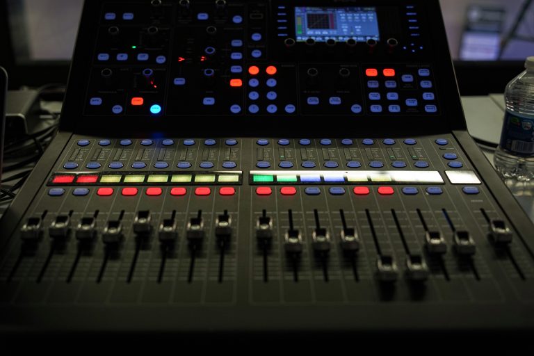 A close-up of a professional audio mixing console with various sliders, buttons, and a digital display screen, illuminated by colored lights. There is a water bottle to the side.