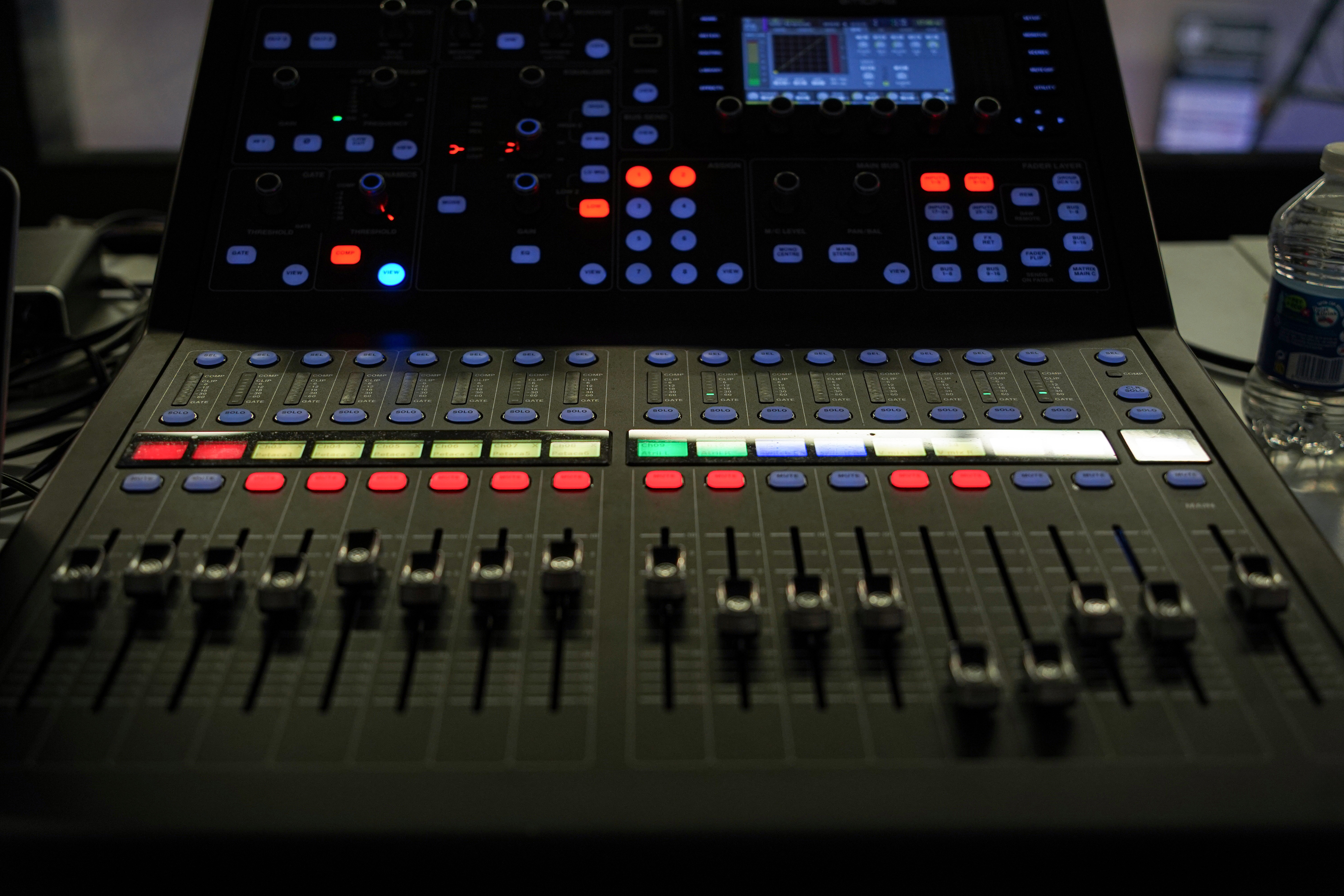 A close-up of a professional audio mixing console with various sliders, buttons, and a digital display screen, illuminated by colored lights. There is a water bottle to the side.