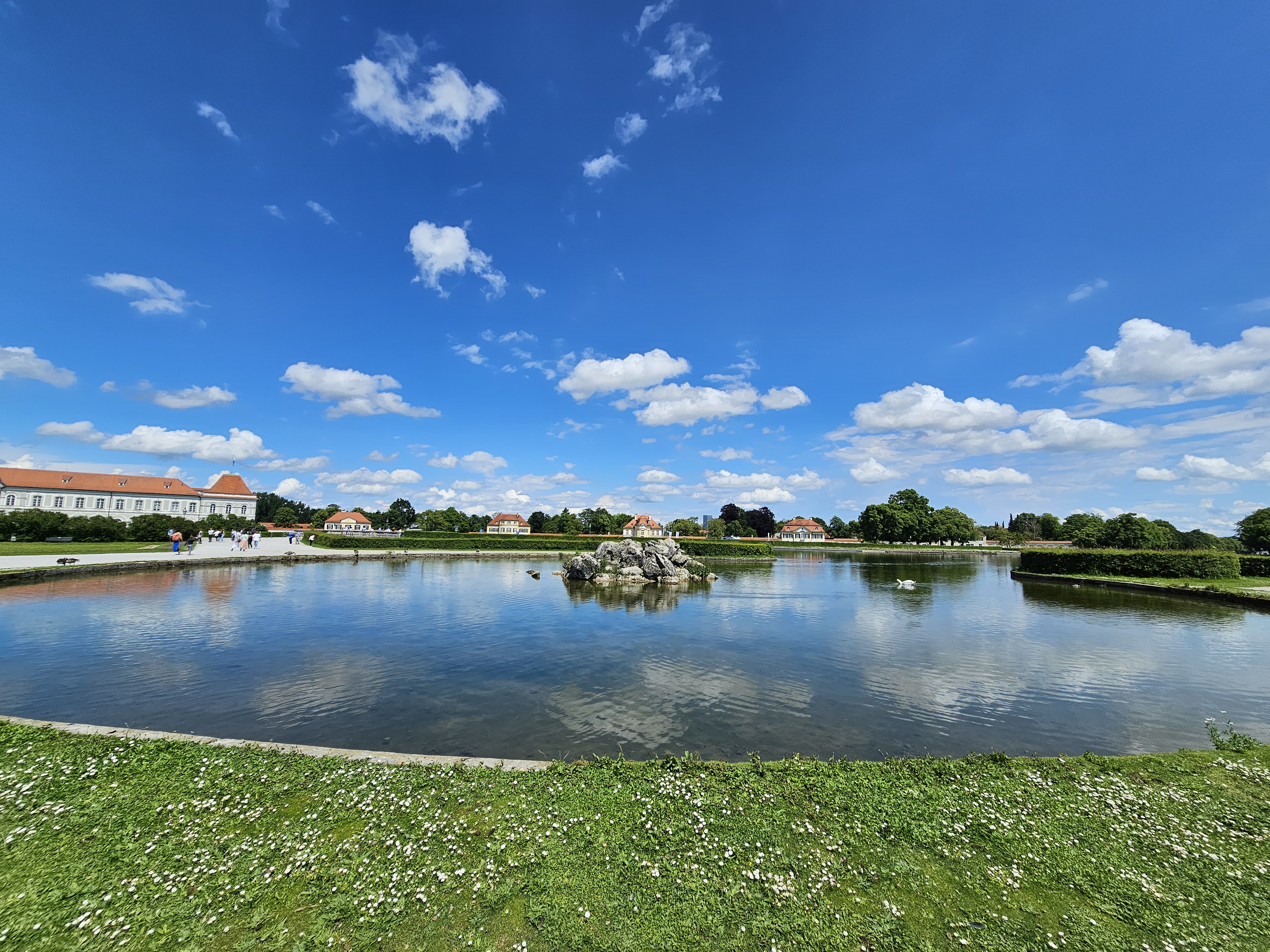 The Nymphenburg Palace reflected in the water on a sunny day with a blue sky and scattered clouds. The palace is located on the left side of the image, and its reflection is visible in the calm water. A small fountain is positioned in the center of the water, and the surrounding landscape includes green grass and trees.