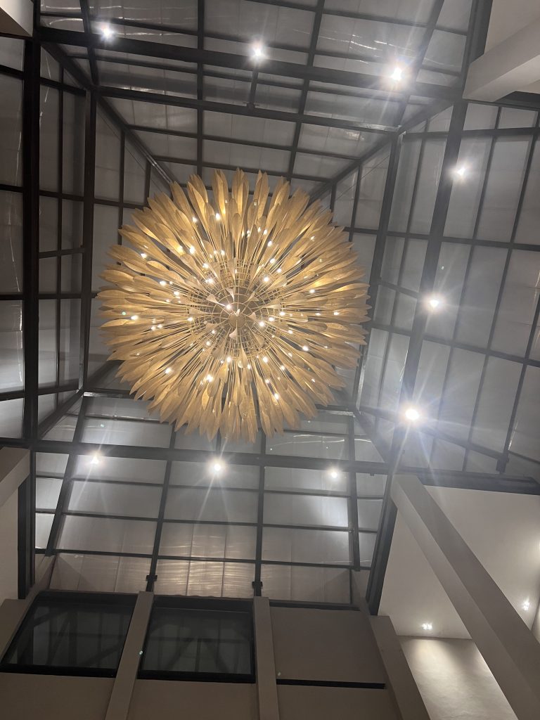 A large, intricate chandelier with gold leaf-like structures is suspended from a high ceiling with a grid of metal beams and glass panels. The chandelier emits a warm glow, illuminating the space below.