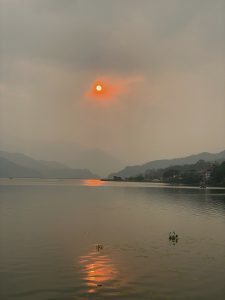 A serene lake scene at sunset with the sun glowing bright orange, partially obscured by clouds. The sun's reflection creates a shimmering path on the water. The distant shoreline features silhouettes of hills and trees, with a few scattered buildings visible.