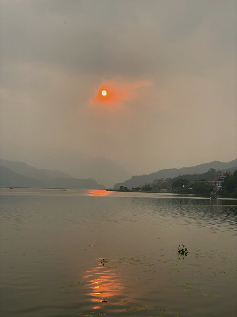 A serene lake scene at sunset with the sun glowing bright orange, partially obscured by clouds. The sun’s reflection creates a shimmering path on the water. The distant shoreline features silhouettes of hills and trees, with a few scattered buildings visible.